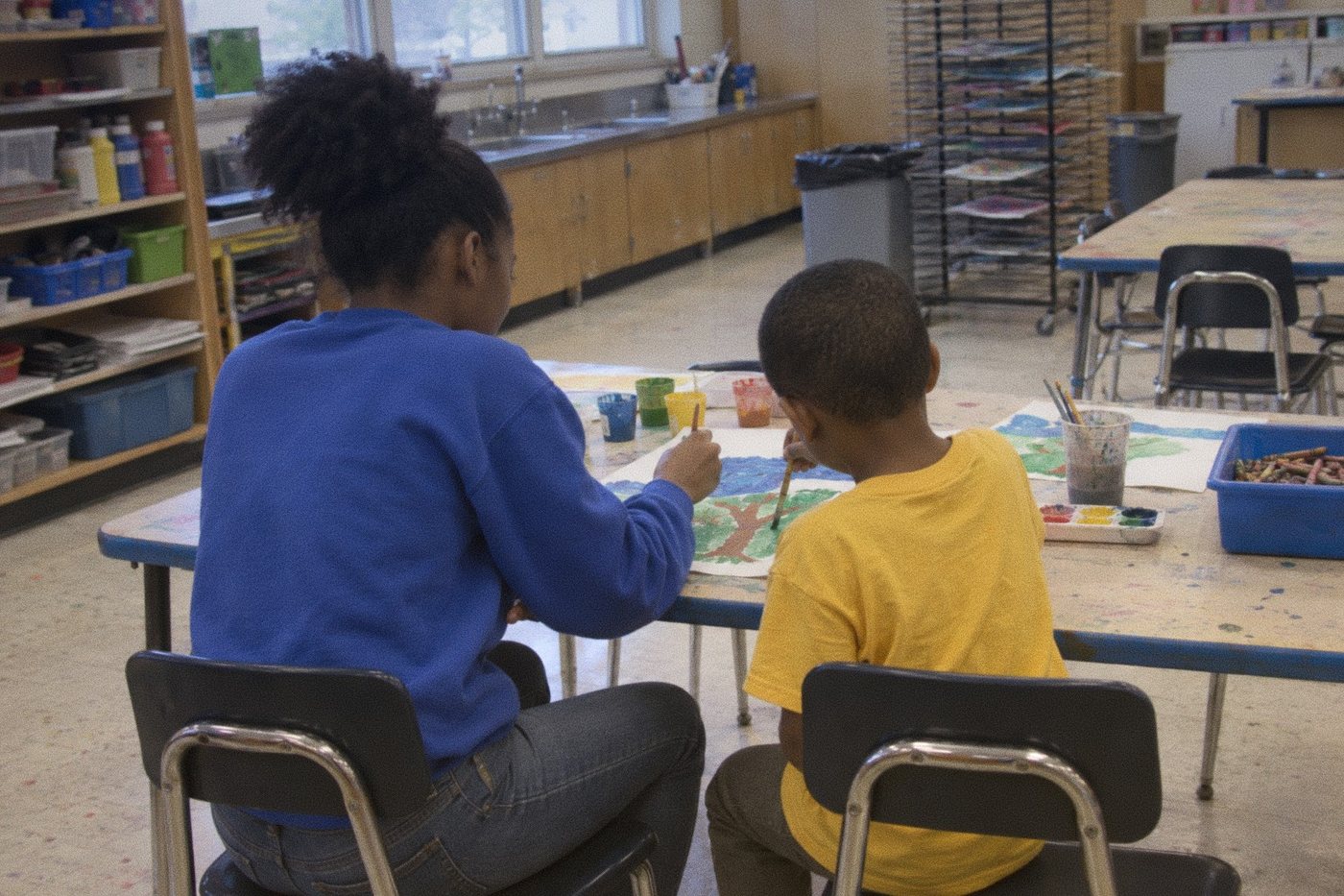 A teenage Black girl in a blue sweatshirt helping a younger Black boy in a yellow t-shirt paint a picture at an art room table, seen from behind